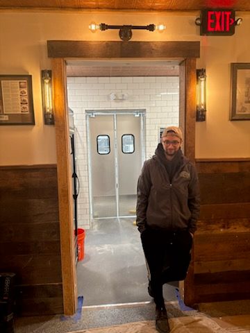 A crew member standing in the finished doorway frame during build-out, unfinished tile hallway behind him